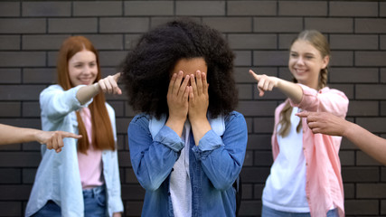 Humiliated black schoolgirl covering face hands, college peers pointing fingers