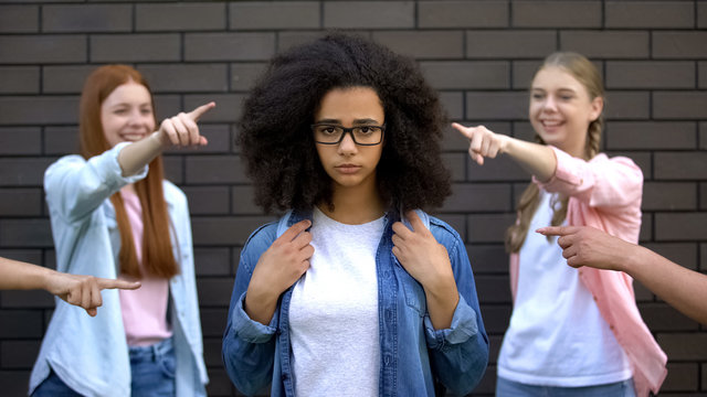 Mocking classmates pointing fingers at black female teenager in eyeglasses