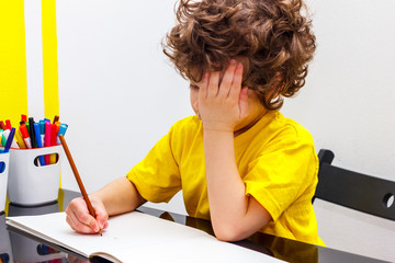 Tired little boy sitting at table, with hishead on his arm, writing in her notebook while doing homework.