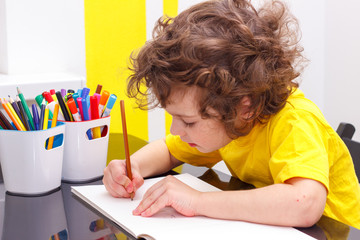 Lovely curly-headed boy in yellow t-shirt writes copy-books, letters