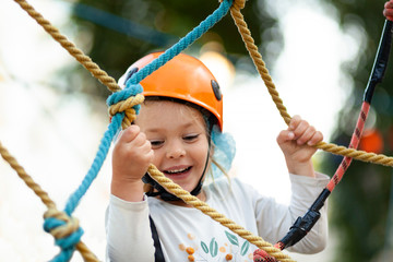 Little girl in helmet climbs ropes in adventure park outdoors. Extreme sport, active leisure on nature.