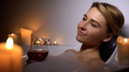 Attractive woman lying in bath with foam bubbles and candles, drinking wine