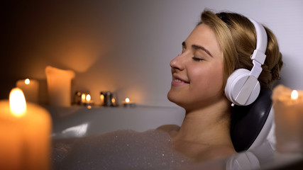 Positive girl in headset lying in bath with foam and candles, listening to music
