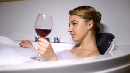 Pretty female with glass of red wine resting in bath with foam bubbles, pleasure