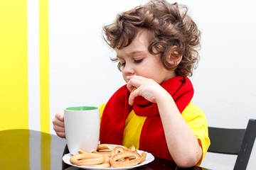 Little curly boy drinking from cup of hot drink tea in cold weather, sitting at home in scarf in kitchen at the table