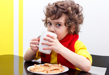 Little curly boy drinking from cup of hot drink tea in cold weather, sitting at home in scarf in kitchen at the table