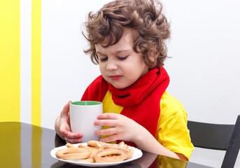 Little curly boy drinking from cup of hot drink tea in cold weather, sitting at home in scarf in kitchen at the table