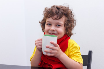 Little curly boy drinking from cup of hot drink tea in cold weather, sitting at home in scarf in kitchen at the table