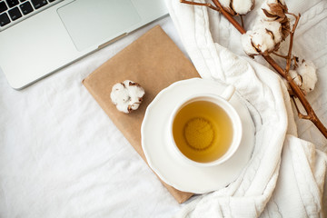 A white mug with tea, cotton, a laptop, a plaid and a book stand on the bed. Breakfast in bed. Cozy. Autumn. Winter.