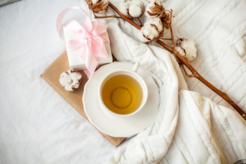 White mug with tea, cotton, plaid, book and white gift box with a pink ribbon on the bed. Breakfast in bed. Cozy. Autumn. Winter.