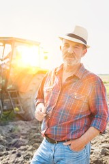 Portrait of senior farmer standing in field while holding wheat crop with yellow lens flare in...