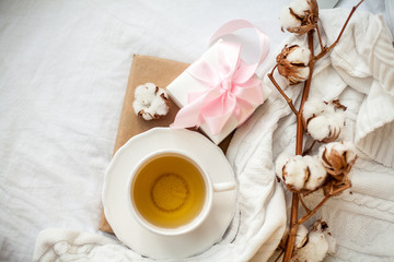 White mug with tea, cotton, plaid, book and white gift box with a pink ribbon on the bed. Breakfast in bed. Cozy. Autumn. Winter.