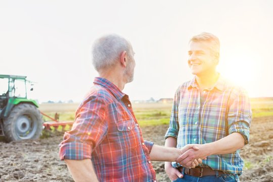 Mature farmer shaking hands with senior farmer in field with yellow lens flare in background