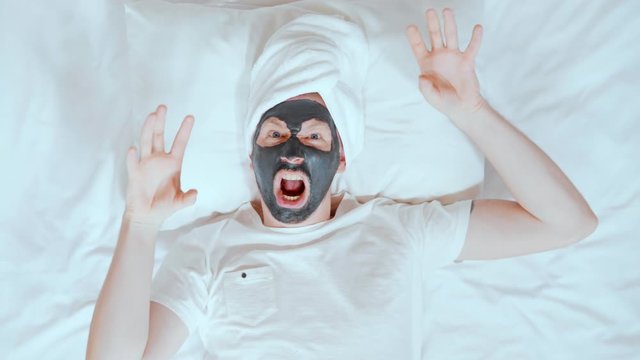 man with a white towel on his head and black cosmetic mask lying on the bed have fun at home