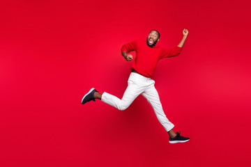 Fototapeta na wymiar Full length photo of cheerful guy jumping raising hands screaming wearing pullover isolated over red background