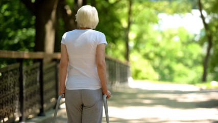 Elderly woman moving with walking frame at rehabilitation center park, back view