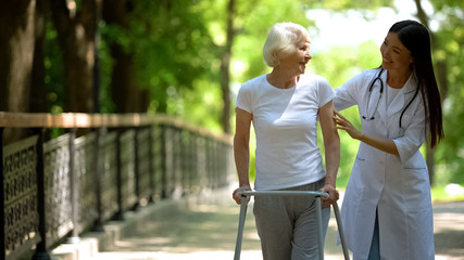 Female doctor helping elderly woman with walking frame, hospital park, outdoors