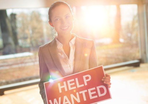 Confident young businesswoman holding red help wanted sign while standing at office with lens flare in background