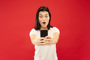 Naklejka premium Caucasian young woman's half-length portrait on red studio background. Beautiful female model in white shirt. Concept of human emotions, facial expression. Making selfie or her own vlog, blog.