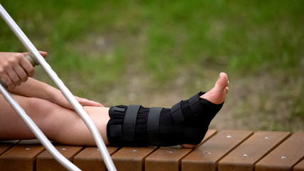 Female with crutch sitting bench with ankle brace on leg, bone fracture, strain - obrazy, fototapety, plakaty