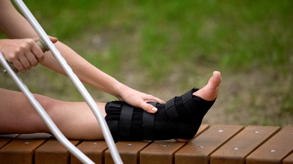 Woman with ankle brace holding crutch sitting outdoors, trauma bandage, health