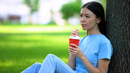 Happy female drinking lemonade through straw sitting garden tree, refreshment