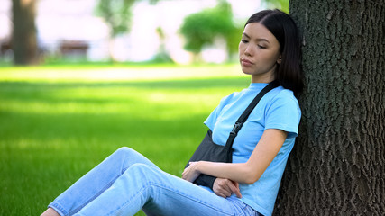 Sad lady with injured arm in supporting sling sitting tree in park, medicine