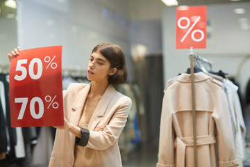 Waist up portrait of beautiful woman hanging red SALE signs on window display in clothes store,...