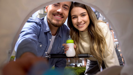 Smiling parents putting baby pacifier to carriage, mum holding milk bottle