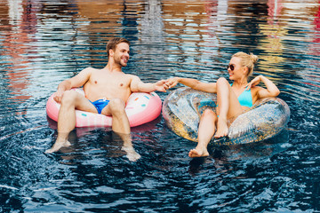 smiling couple holding hands and looking at each other while lying on swim rings in pool