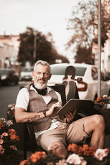 Adult businessman work in cafe on the city street. Stylish bearded man write note in notebook or laptop. Male model portrait outdoors on buildings background