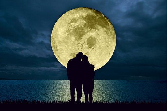 Silhouette of couple embracing standing at the beach at night watching the moonlight from a big full moon shining in the ocean. Love and romance concept. Moon image furnished by NASA.