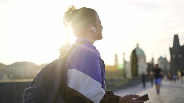Young sportive girl walking in historical centre of Prague at morning time. Side view woman 80's style clothes moving with smartphone, she enjoying music in headset and happily dancing in backlit