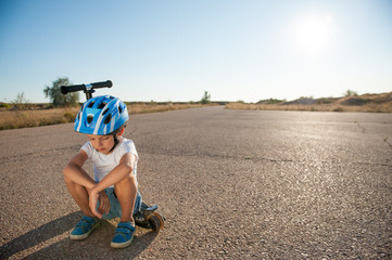 sad little caucasian boy in helmet sitting on his scooter on asphalt empty road during summer hot sunset in California with copy space