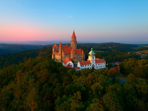 Aerial view on a romantic fairytale castle on a knoll in highland landscape, surrounded by trees. Gothic castle with high towers, red roofs, stone walls. Bouzov castle, Moravian region, Czechia.