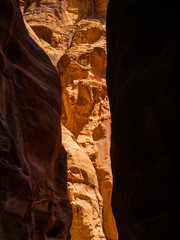 Steep cliffs of a canyon leading to the historical site of Petra, Jordan, light shining from blue sky on dark sandstone walls