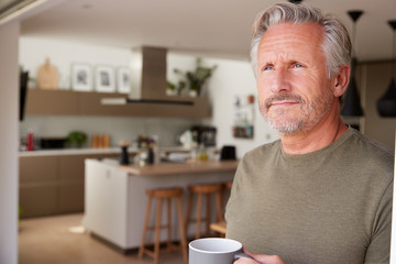 Senior Man Standing And Looking Out Of Kitchen Door Drinking Coffee