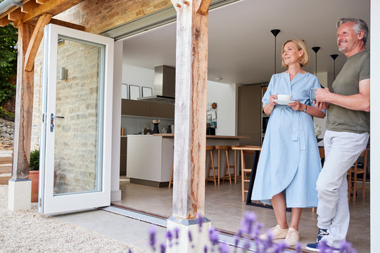 Senior Couple Standing And Looking Out Of Kitchen Door Drinking Coffee