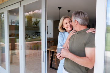 Senior Couple Standing And Looking Out Of Kitchen Door Drinking Coffee