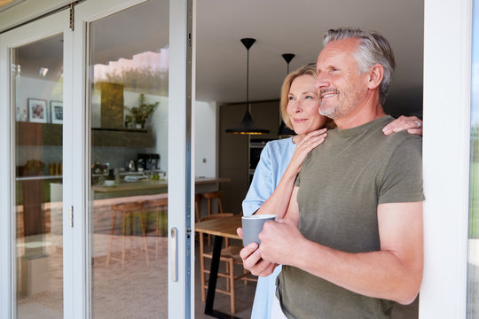 Senior Couple Standing And Looking Out Of Kitchen Door Drinking Coffee