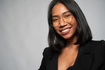 Portrait of attractive young Asian smiling woman in elegant black blazer and glasses posing over light gray wall background. Mock up copy space