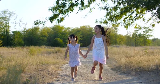 Two pretty beautiful little girls four and seven years old in white summer dresses,  skipping along a dusty country trail holding hands on warm summer evening. Slow motion 4k 50 fps