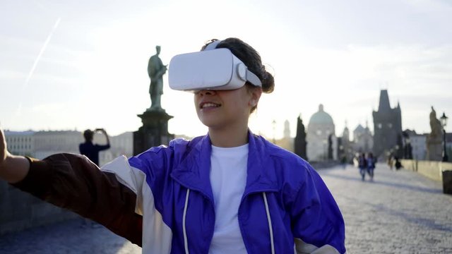 Woman 80's style clothes walking on famous Charles bridge in Prague. Girl wearing VR headset, moving hands before itself, touching virtual objects at defocused background old European town