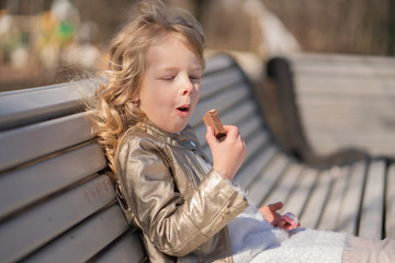 emotional portrait of a little girl with sweets, sitting on the bench in the city park outdoor alone. pretty child have a bad tasty snack.