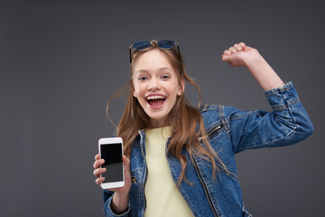 Happy teen girl in jean jacket holding modern cellphone and raising hand