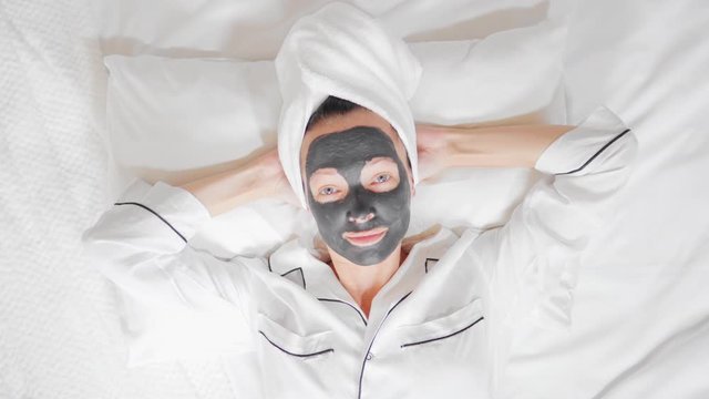 woman with a white towel on her head and black cosmetic mask on the bed relaxing at home wellness, spa, skin care