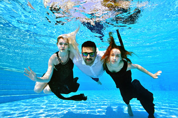 Two young girls, a blonde and a redhead, swim and pose underwater embracing a guy of Eastern nationality in sunglasses. Girls in dresses, a guy in a white shirt. Fashion portrait