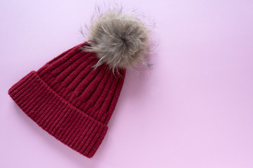 Cozy and warm winter flat lay with copy space. Dark red knitted hat with fur pompom on pink background