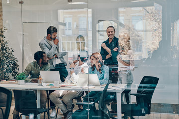 Group of young modern people in smart casual wear communicating and using modern technologies while working in the office