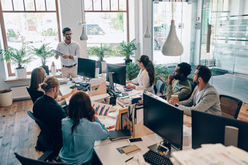 Group of young modern people in smart casual wear communicating and using modern technologies while working in the office